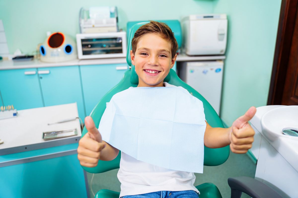 Your Child’s First Cavity Young child giving thumbs up in dental office wearing bib, smiling at camera.