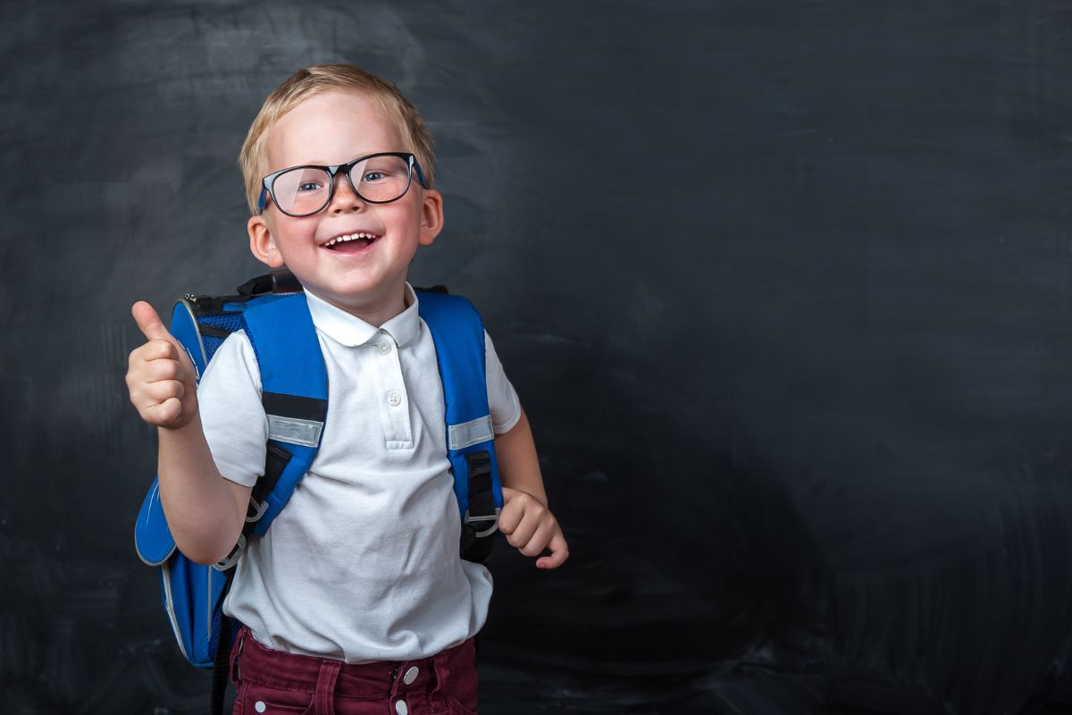 Is Your Child’s Smile Ready for School Young boy wearing glasses and blue backpack giving thumbs up against dark chalkboard background.