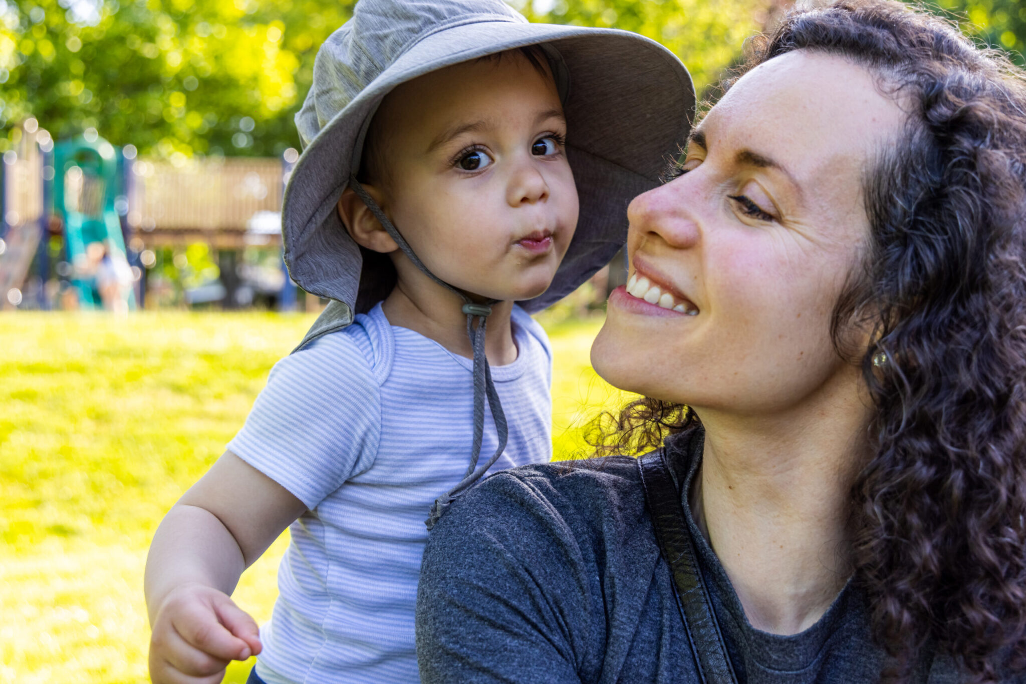 Smiling mother holding a young child in a sun hat at a park near Long Island Pediatric Dentistry in Lawrence, NY