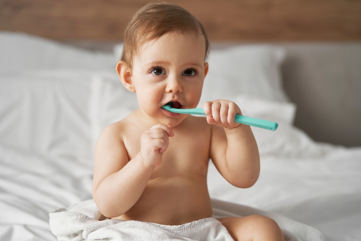 Baby sitting on a bed, chewing on a teal toothbrush with a towel draped around the waist.