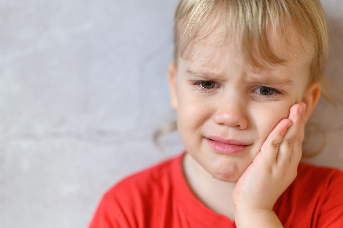 kids emergency Crying toddler in a red shirt holding their cheek, with tears on their face and a pained expression.