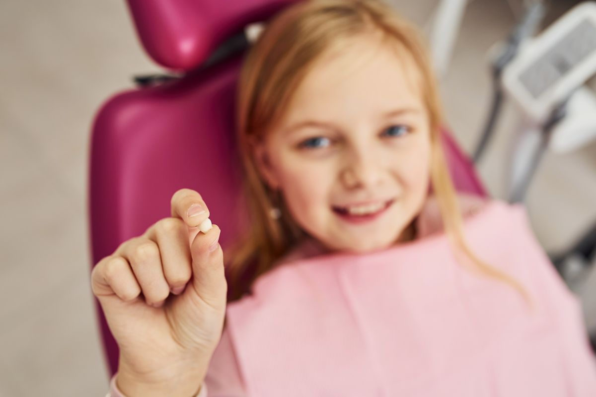 Smiling child in a dentist’s chair holding a recently lost baby tooth between fingers.