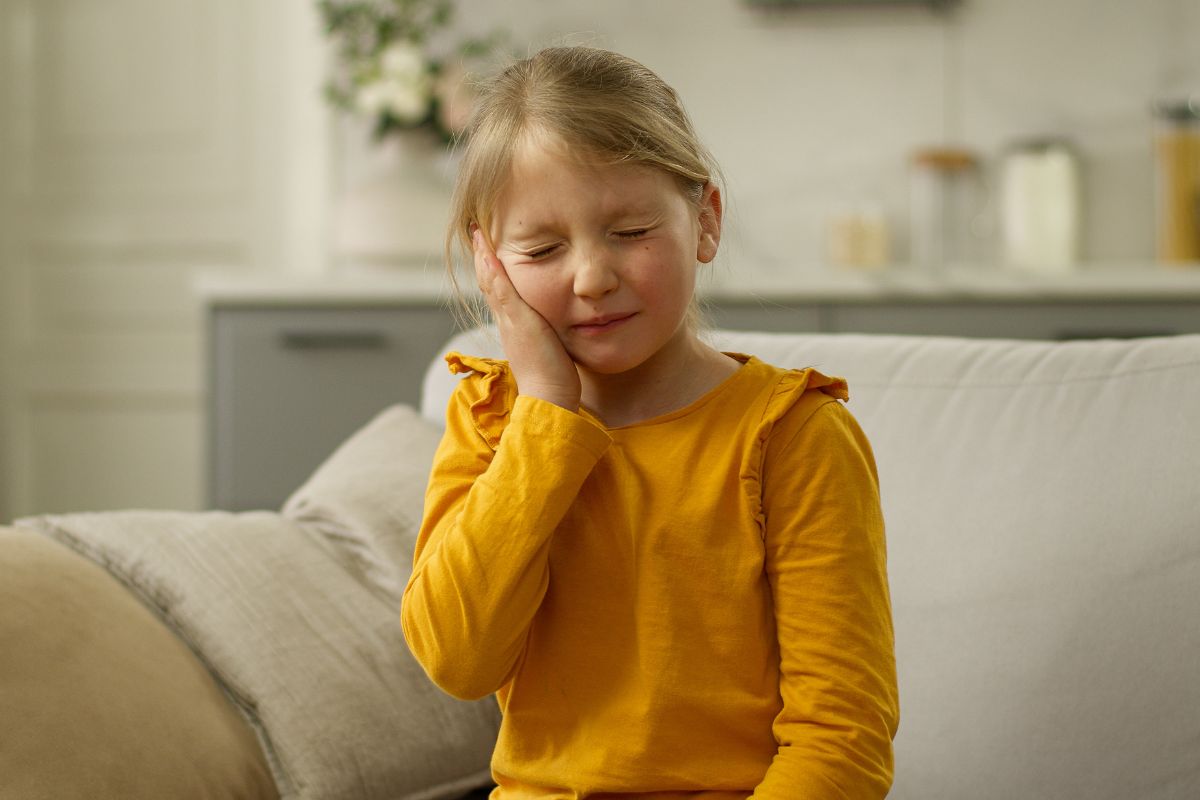 A young girl sits at home holding her cheek, showing signs of dental discomfort that may need attention during school hours.